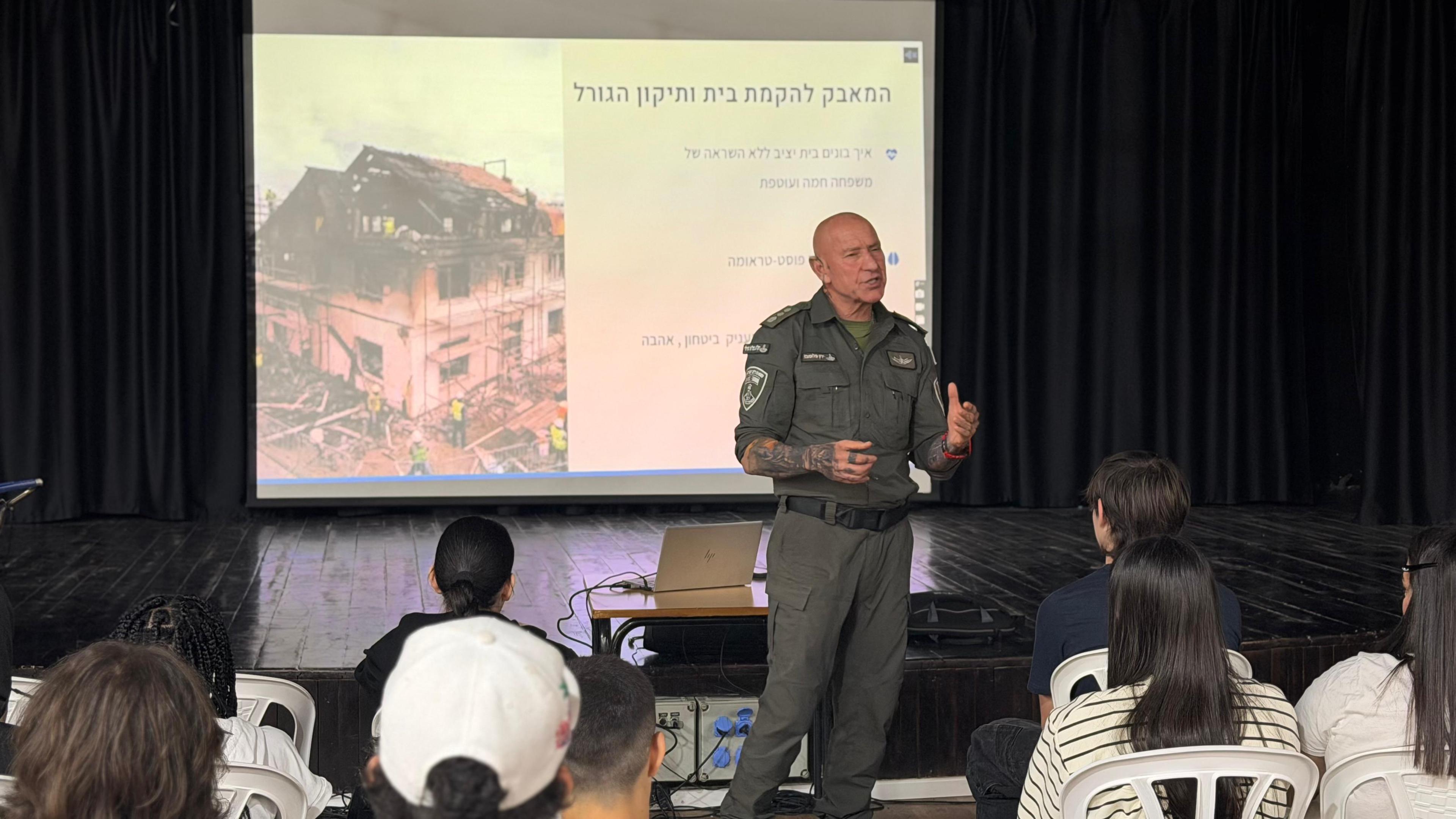 ירון פלומבו מרצה בתיכון יוצר עתיד, מצגת "המאבק להקמת בית ותיקון הגורל" ברקע